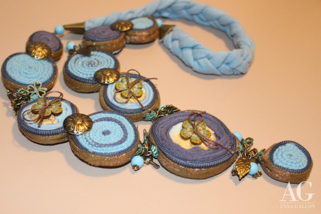 Collana realizzata interamente a mano nei toni dell'azzurro, giallo e bronzo, utilizzando stoffe, passamaneria, perline ciondoli in metallo e fiori in legno.